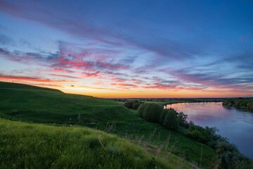 Fototapeta na wymiar Summer landscape with a river flowing between green hills against the backdrop of a colorful sunset sky. An epic evening landscape with a fantastic sunset.