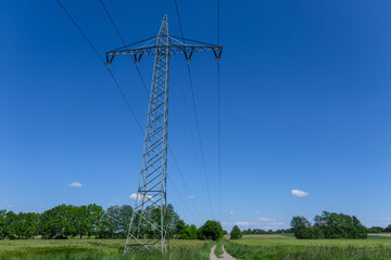 Obraz premium Konzept Energie: Strommast mit Überleitungen in grüner Natur neben einem Feldweg und bei blauem Himmel