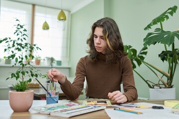 Talented creative guy teenager painting with watercolors, sitting at the table at home