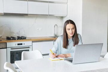 Obraz na płótnie Canvas Executive freelance woman using laptop for remote work from home. Female student in casual wear sits at the desk in the kitchen, watching online webinars or training and taking notes, writes down
