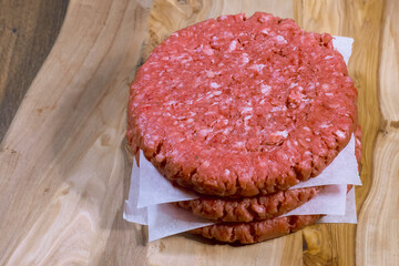 Stack of three fresh uncooked beef burgers separated with cooking paper on a cutting wooden board and table. Food industry.