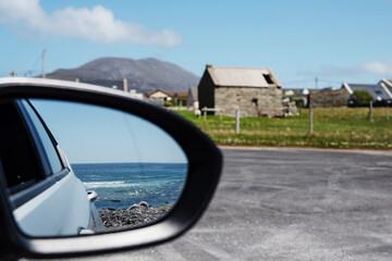 Obraz premium Blue ocean surface reflection in a car mirror in focus. Small town and a mountain out of focus in the background. Travel and tourism concept. Achill island, county Mayo, Ireland. Warm day