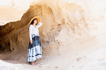 Naklejka premium woman in a cave on the beach looking at the sea, wears long dress and straw hat