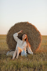 Naklejka na ściany i meble young woman in the beautiful light of the summer sunset in a field is sitting near the straw bales. beautiful romantic girl with long hair outdoors in field