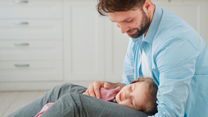 Smiling dad with beard looks and caresses small girl child with long fair hair sleeping on father knees