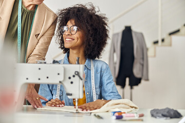 Woman boss checks work of African-American designer at sewing machine in fashion studio
