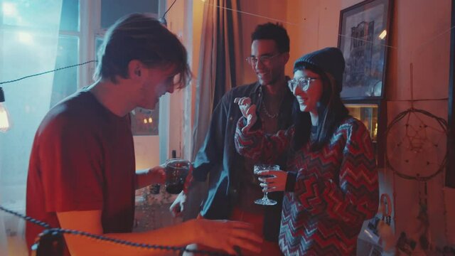 Company of young tipsy friends chatting, smoking and dancing with drinks at home retro party in room decorated with garland