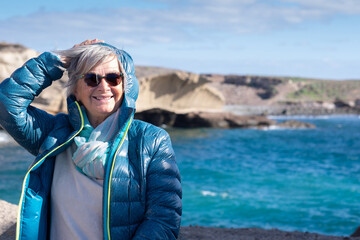 Portrait of senior woman  at sea in windy day, standing on the cliff. Joyful moment appreciating freedom and beauty in nature