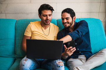 Smiling ethnic gays sharing laptop and smartphone on sofa