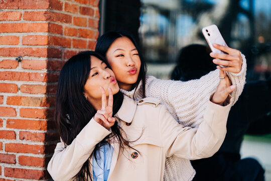 Happy young Asian ladies pouting lips while taking selfie on street