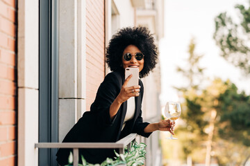 Positive black woman in stylish outfit taking selfie on balcony