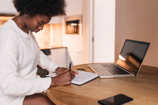 Focused ethnic female freelancer taking notes at table with laptop