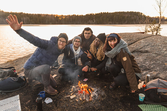 Cheerful diverse friends sitting around bonfire on river bank
