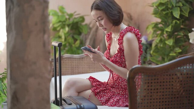 happy asian tourist woman with suitcase using application on mobile smart phone booking flight ticket online in summer tropical hotel room. online purchasing on internet. mobile smartphone application