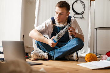 Young man using laptop and sewing machine while working on craft rug