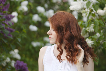 A beautiful young woman with red curly hair and freckels in a white dress in a white lilac garden in spring time.