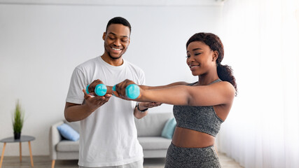 Joyful black guy helping his girlfriend exercise with dumbbells in living room, banner design