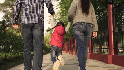 Dad, daughter, mom, are playing in city park, happy child is holding parents' hands and jumping. Teamwork. Happy family runs in park on street, holding hands in spring, summer. Happy healthy childhood