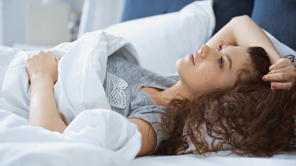 young curly woman looking up while lying on bed in morning