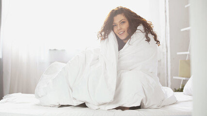 cheerful young woman wrapped in white blanket sitting on bed