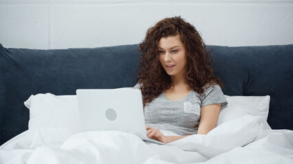 curly freelancer smiling while using laptop in bed
