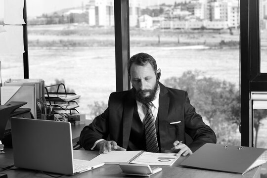 Black and white image of Bearded man wearing shirt tie and black suit sits in office at table, holds pen in his hand looking at us. Man with documents and folders working with laptop at desk in office
