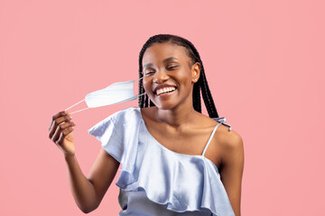 Coronavirus is over. Happy black woman with afro bunches taking off her face mask on pink studio...