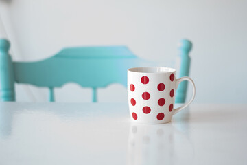 A large white cup of coffee or tea with red dots stands on a white table. A navy blue Greek-style chair stands next to the table. Illuminated kitchen