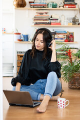 Smiling female businesswomen with headphones is siting on the floor watches webinar, working from home. Looking at her laptop computer wearing formal wear using earphones.
