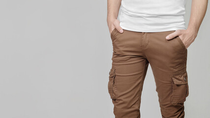 Model wearing brown cargo pants with hands in pockets on grey background, empty place for text on the left. Mockup, template. - obrazy, fototapety, plakaty