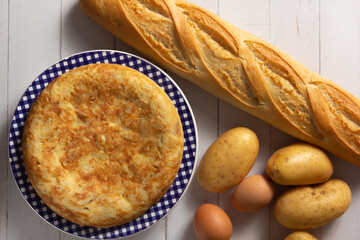 Tortilla de Patatas española. Delicioso plato salado basado en Huevo, Patatas y a veces Cebolla. Acompañada por patatas, una barra de pan, y huevos. En fondo de madera blanca, entera y sin cortar. 