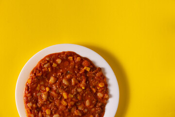 Traditional Hot chili sauce. Chopped meat, white beans, korn pepper, paprika in a tomato sauce.  on a white plate and a yellow background