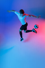High angle view of young man, male soccer football player training isolated on gradient blue pink background in neon light.