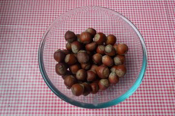Hazelnuts lie in a transparent bowl on the table. On the table is a pink checkered tablecloth. View from above.