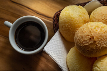 Uma xícara branca cheia de café e uma pequena cesta de vime com pães de queijo sobre uma superfície de madeira.