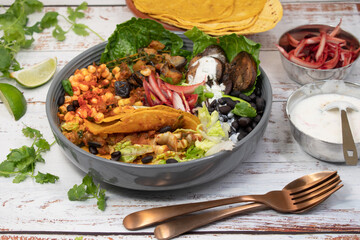 Vegan Mexican taco bowl consisting of spicy corn, black beans, eggplant and mushroom, salad, pickles. White wooden table