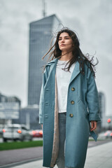 Portrait of a business woman concept. Isolated photo of the bru young girl in overcoat with skyscrape behind her in the center of the modern city. Woman is standing in front of modern office buildings
