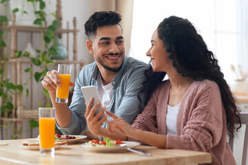 Joyful Arab Couple Eating Breakfast Together And Using Smartphone In Kitchen