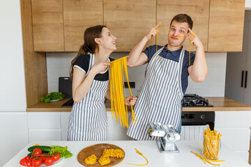 Young couple in aprons having fun with pasta noodles. Family cooking italian vegan food at home....