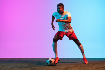 Portrait of African professional football player training isolated on gradient blue pink background...