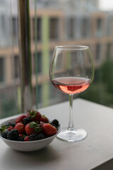 A glass of rose wine is on the table on the balcony, next to a plate of berries. Drink wine and eat berries. The mood of summer. Wine with fruit. Summer vacation with wine. Alcoholic beverages.