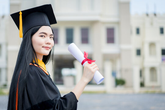 A young beautiful Asian woman university graduate in graduation gown and mortarboard holds a degree certificate stands in front of the university building after participating in college commencement