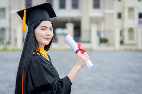 A young beautiful Asian woman university graduate in graduation gown and mortarboard holds a degree certificate stands in front of the university building after participating in college commencement