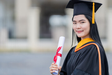 A young beautiful Asian woman university graduate in graduation gown and mortarboard holds a degree...