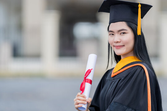A young beautiful Asian woman university graduate in graduation gown and mortarboard holds a degree certificate stands in front of the university building after participating in college commencement