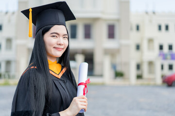 A young beautiful Asian woman university graduate in graduation gown and mortarboard holds a degree...