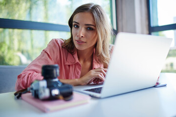 Portrait of attractive female photographer journalist with brunette hair sitting at coworking desktop with digital netbook computer and blurred vintage camera for working remotely with media files
