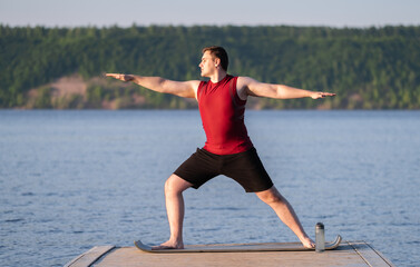 A young man practices yoga on the river bank. The concept of weight loss and sports in nature.