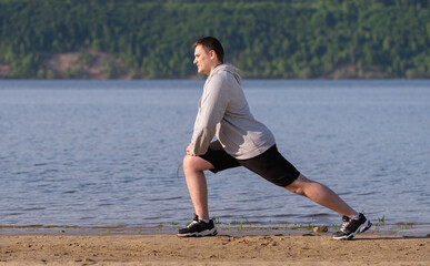 A young man in a gray sweatshirt stretches before jogging on an empty beach at dawn. Slimming and healthy lifestyle concept.