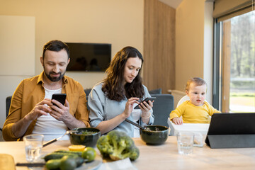 Young parents and their one year baby boy sitting in mobile devices separately during lunch at home. The problem of Internet addiction of parents and raising children